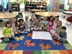 students sitting on floor rug around a gridded posterboard. One holds the book One Hundred Hungry Ants.