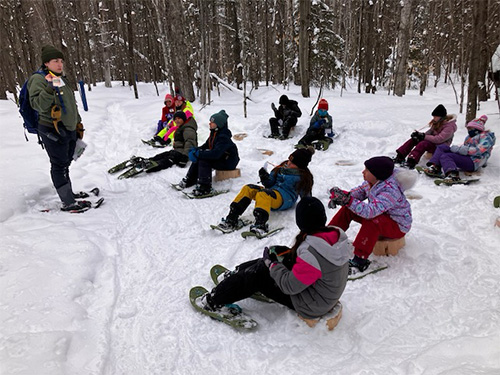 Students sitting on logs in the snow listening to an adult