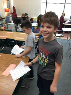 students working at table and closest boy is smiling while holding paper and pencil