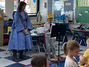 Student with a crown reading to the class next to the teacher dressed as the fairy godmother