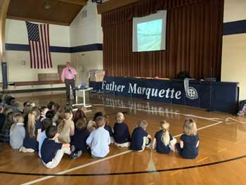 Students sitting on the gym floor listening to an adult who is in front of a Father Marquette sign