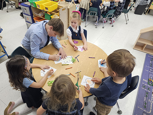 Older student coloring at a round table with pre-k students